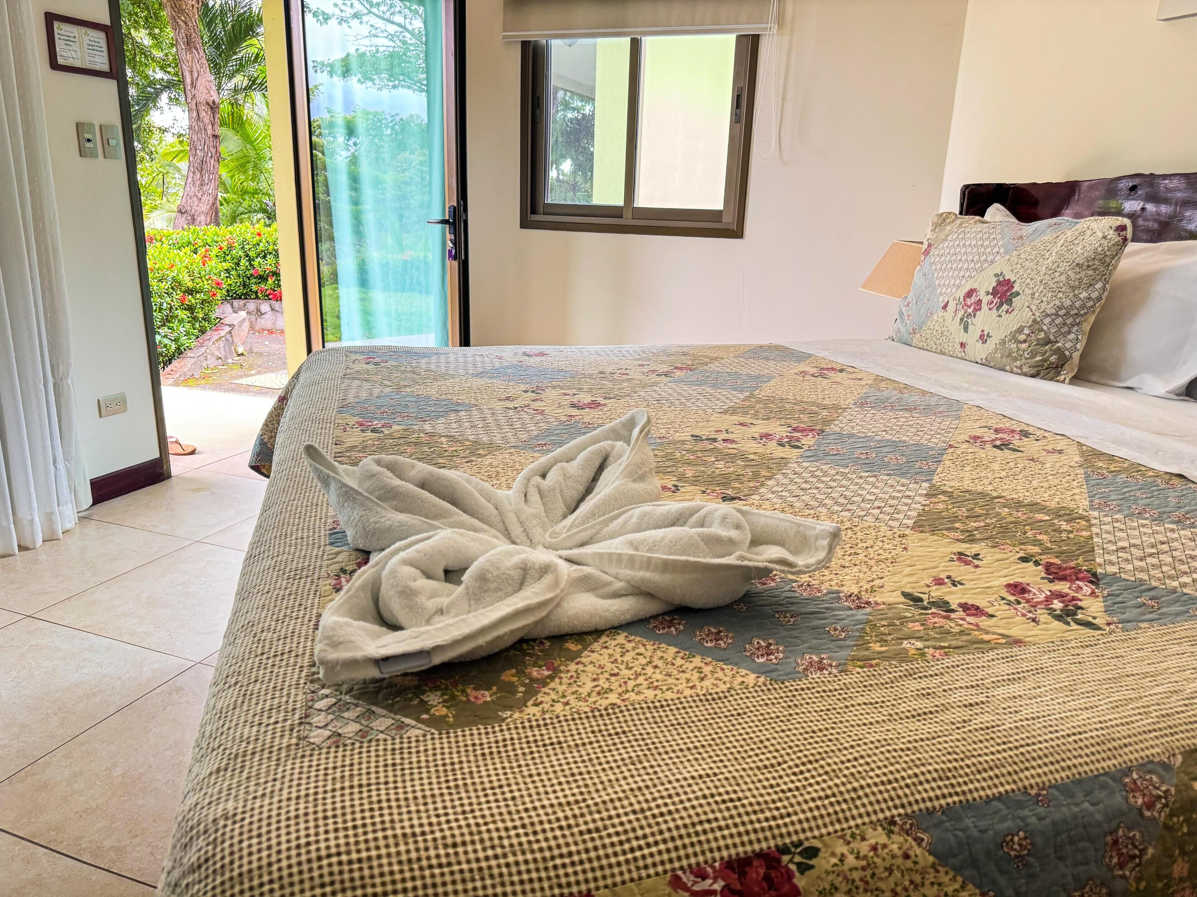 A hotel room with a bed, a window, and a view of greenery outside.