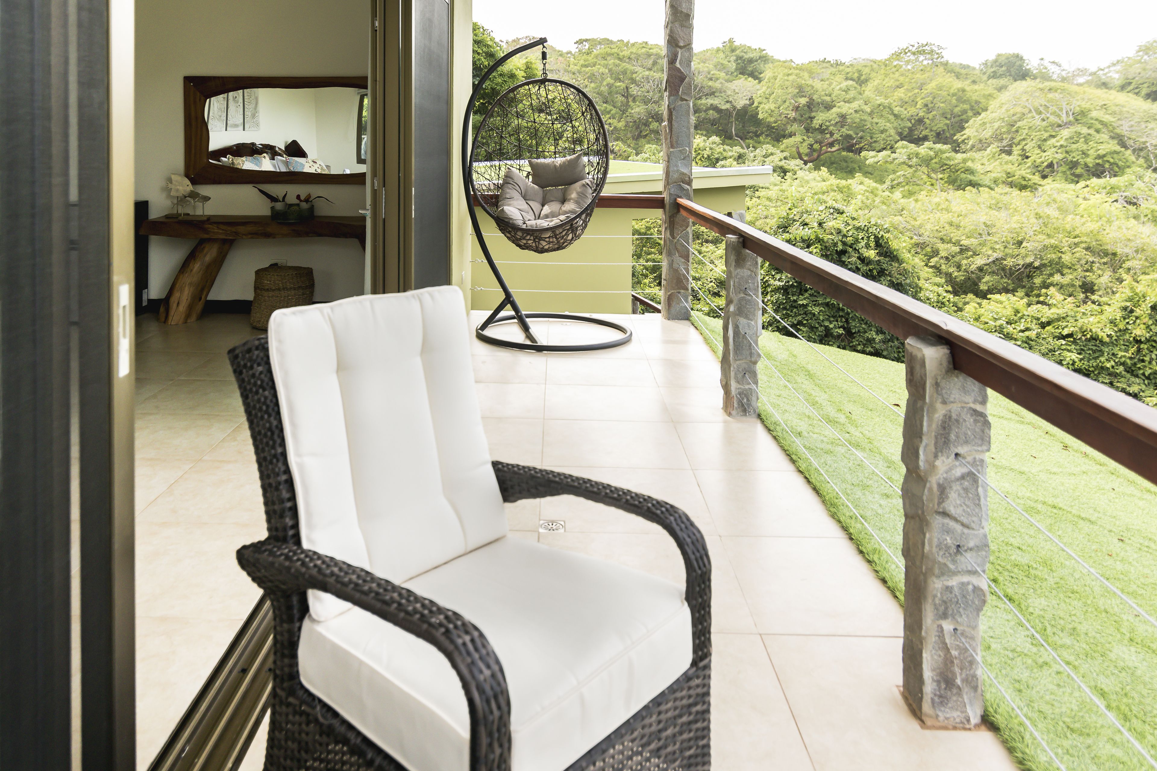 A wicker chair with white cushions on a balcony with a stone railing, overlooking a lush green landscape.