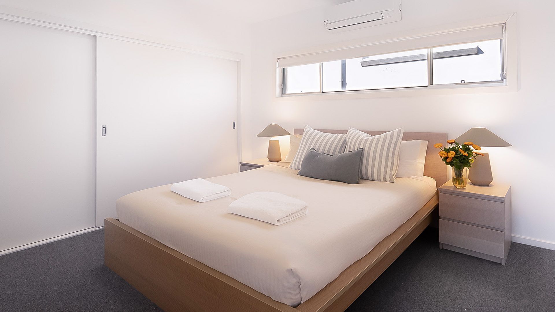 A modern bedroom with a large bed, two bedside tables, a nightstand, a window with blinds, and a ceiling vent.