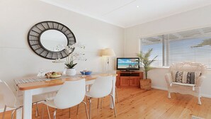 Un salon moderne avec une table à manger en bois, des chaises blanches, un téléviseur et un fauteuil en osier.