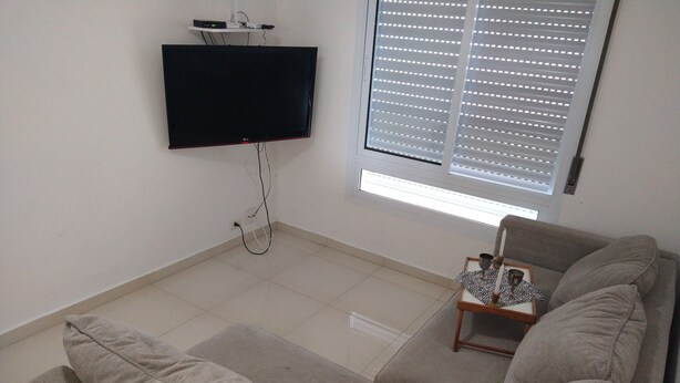 A living room with a flat-screen TV mounted on the wall, a window with blinds, a grey sofa with cushions, and a small table with a tray. - Cozy apartment with garage, Wi-Fi, sea view and only 3 minutes from the beach. (Guaruja)