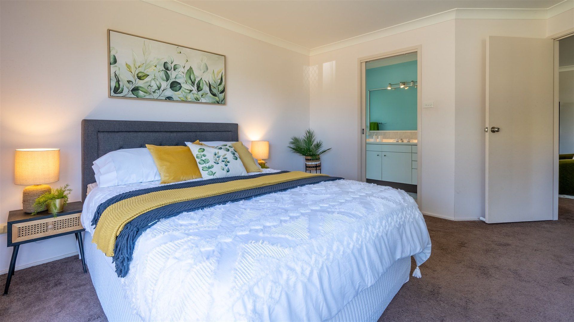 A bedroom with a large bed, bedside lamps, a plant, and a bathroom visible through an open door.
