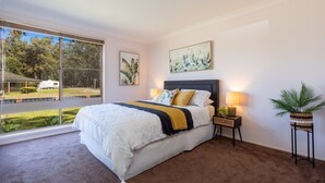 A bedroom with a large window, a bed with white bedding and a yellow throw, a nightstand with a lamp, and a potted plant.