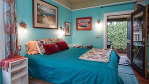 A bedroom with a teal bedspread, a large framed picture, and a small table with a lamp.