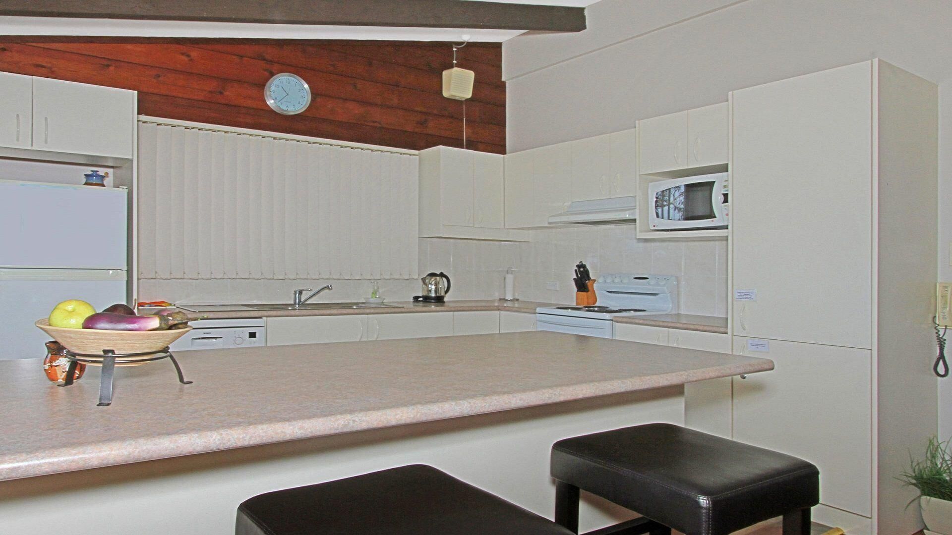Uma cozinha compacta com uma ilha central, armários brancos, um micro-ondas e um relógio na parede.