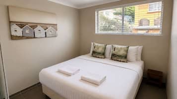 A bedroom with a bed, two pillows, a window with blinds, and a wall-mounted artwork of houses.