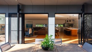 A modern interior with a wooden dining table, a potted plant, and a kitchen area visible through glass doors.