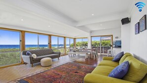 Uma sala ampla com uma grande janela com vista para o mar, uma área de jantar com mesa e cadeiras, e um sofá amarelo com almofadas.
