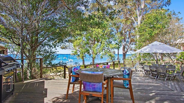 A wooden deck with a table and chairs, overlooking a body of water with boats. - Bay Cottage – stunning water views of Callala Bay (Callala Bay)