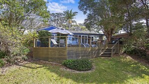 Une terrasse en bois dotée d’un espace salon couvert, d’un escalier et offrant une vue sur une maison avec un toit bleu.
