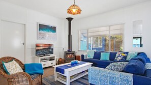 A living room with a television, a sofa, a wicker chair, a table, and a fireplace. - Bay Cottage – stunning water views of Callala Bay (Callala Bay)