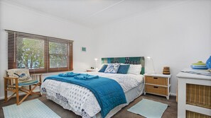 Une chambre à coucher comprenant un lit, des tables de chevet, une chaise, une fenêtre avec des stores et une vue sur la verdure à l’extérieur.