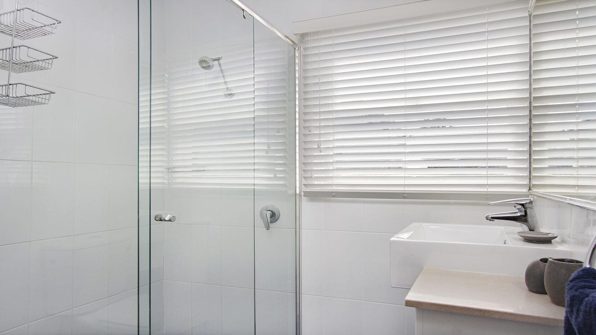 Ein modernes Badezimmer mit einer Duschkabine aus Glas, einem Waschbecken mit Spiegel darüber und einem Fenster mit Jalousien.