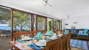A dining area with a wooden table set for six, large windows, and a view of the sea. - Bay Cottage – stunning water views of Callala Bay (Callala Bay)