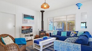 A living room with a television, a fireplace, a sofa, and a wicker chair. - Bay Cottage – stunning water views of Callala Bay (Callala Bay)