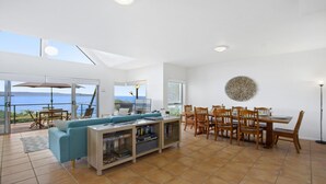 Un vaste espace de vie comprenant un grand canapé, une table à manger avec des chaises et une bibliothèque. De grandes fenêtres offrent une vue sur la mer.
