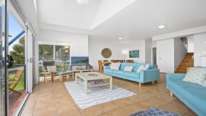 Un salon spacieux avec un canapé bleu, une table basse et un téléviseur. On y trouve également un coin repas avec des chaises et une table, ainsi qu’un escalier menant à un autre niveau.