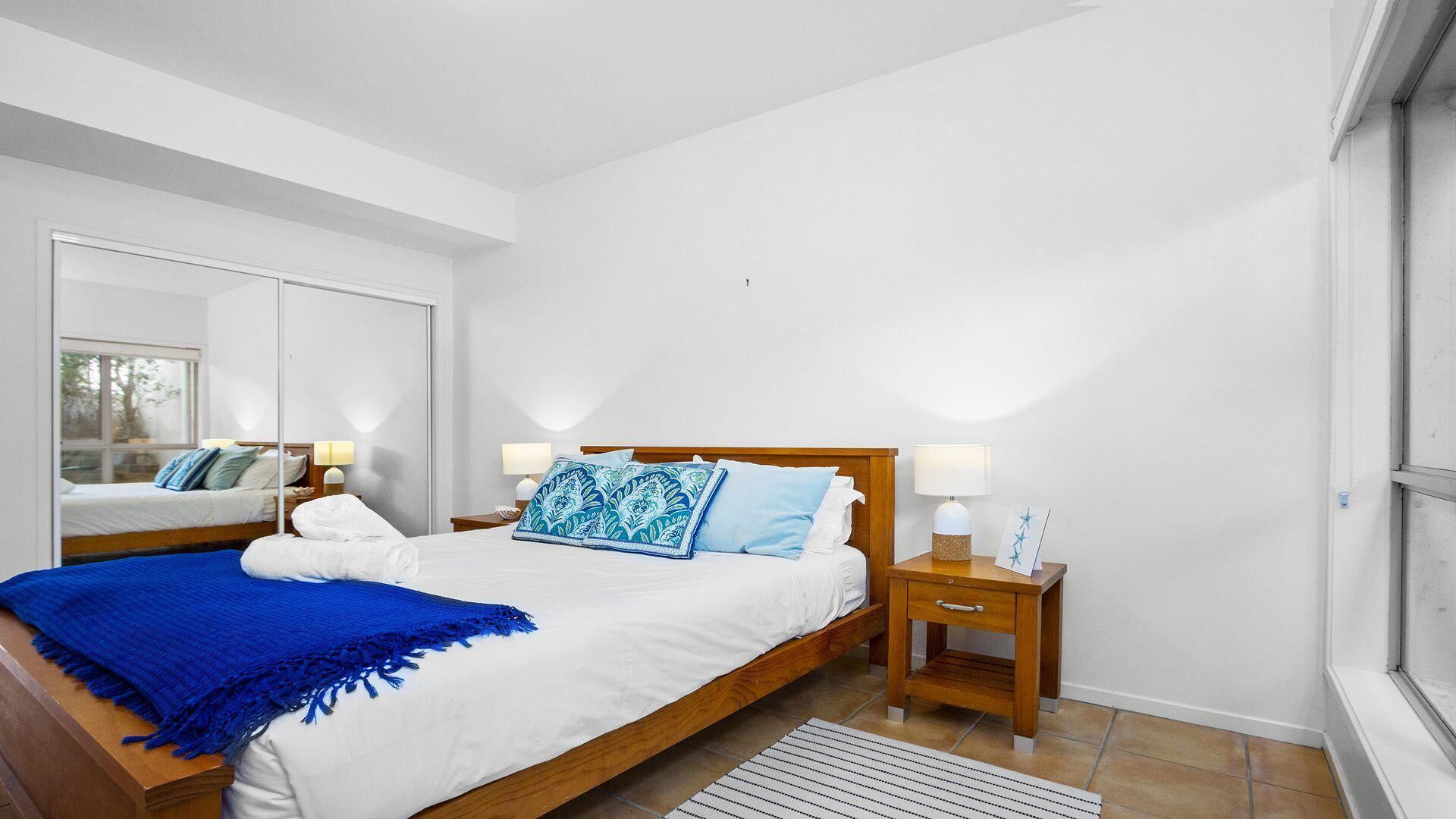 Un dormitorio moderno con una cama de madera, sábanas blancas, un plaid azul y dos mesitas de noche con lámparas.
