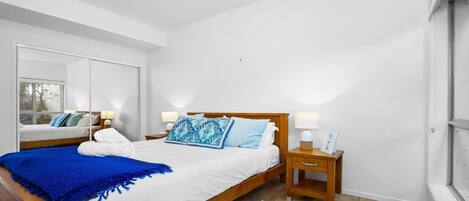Un dormitorio moderno con una cama de madera, sábanas blancas, un plaid azul y dos mesitas de noche con lámparas.