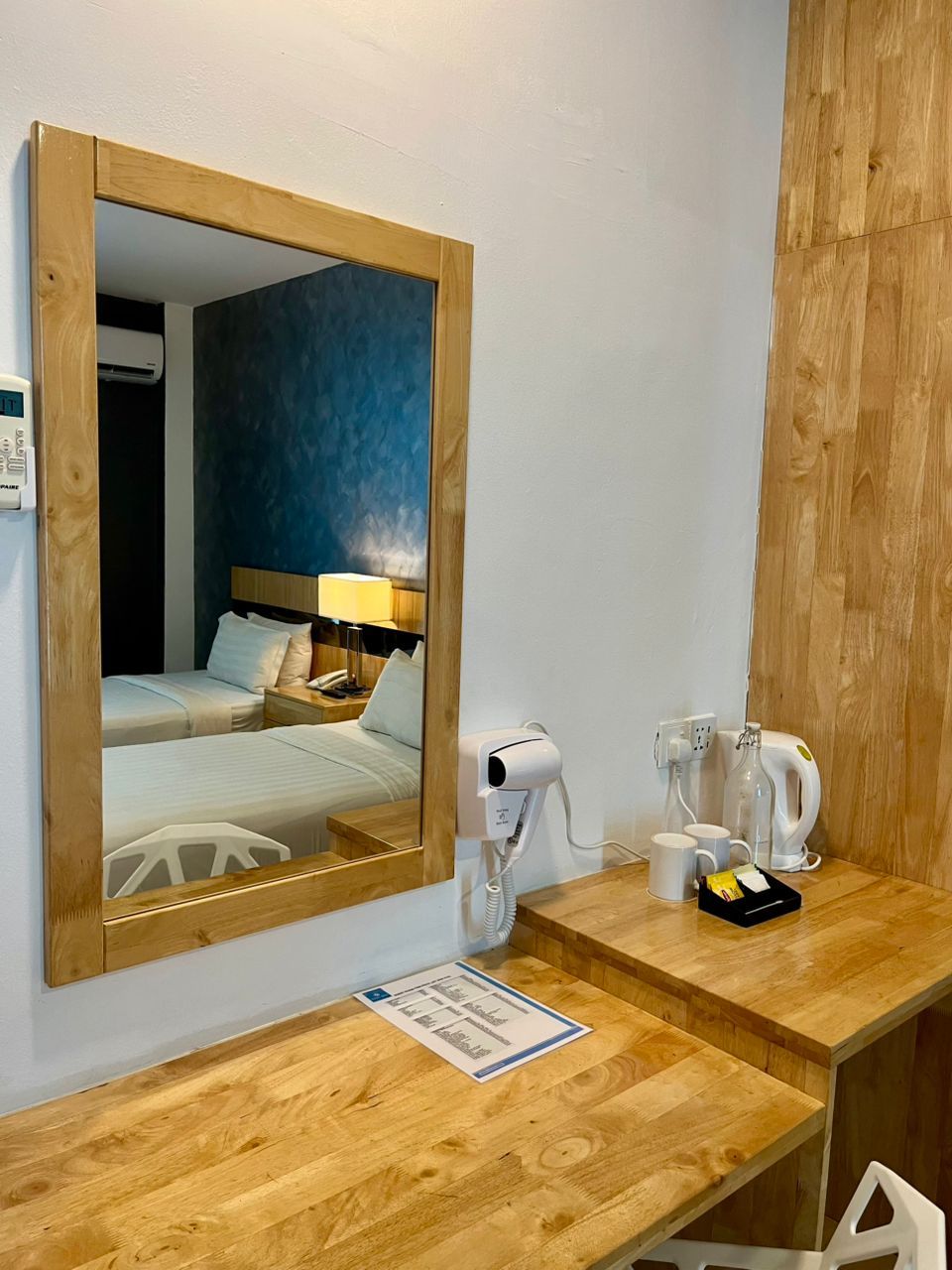 A hotel room with a wooden desk, a hairdryer, a kettle, and a mirror reflecting a bed with a nightstand and lamp.