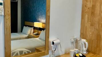 A hotel room with a wooden desk, a hairdryer, a kettle, and a mirror reflecting a bed with a nightstand and lamp.