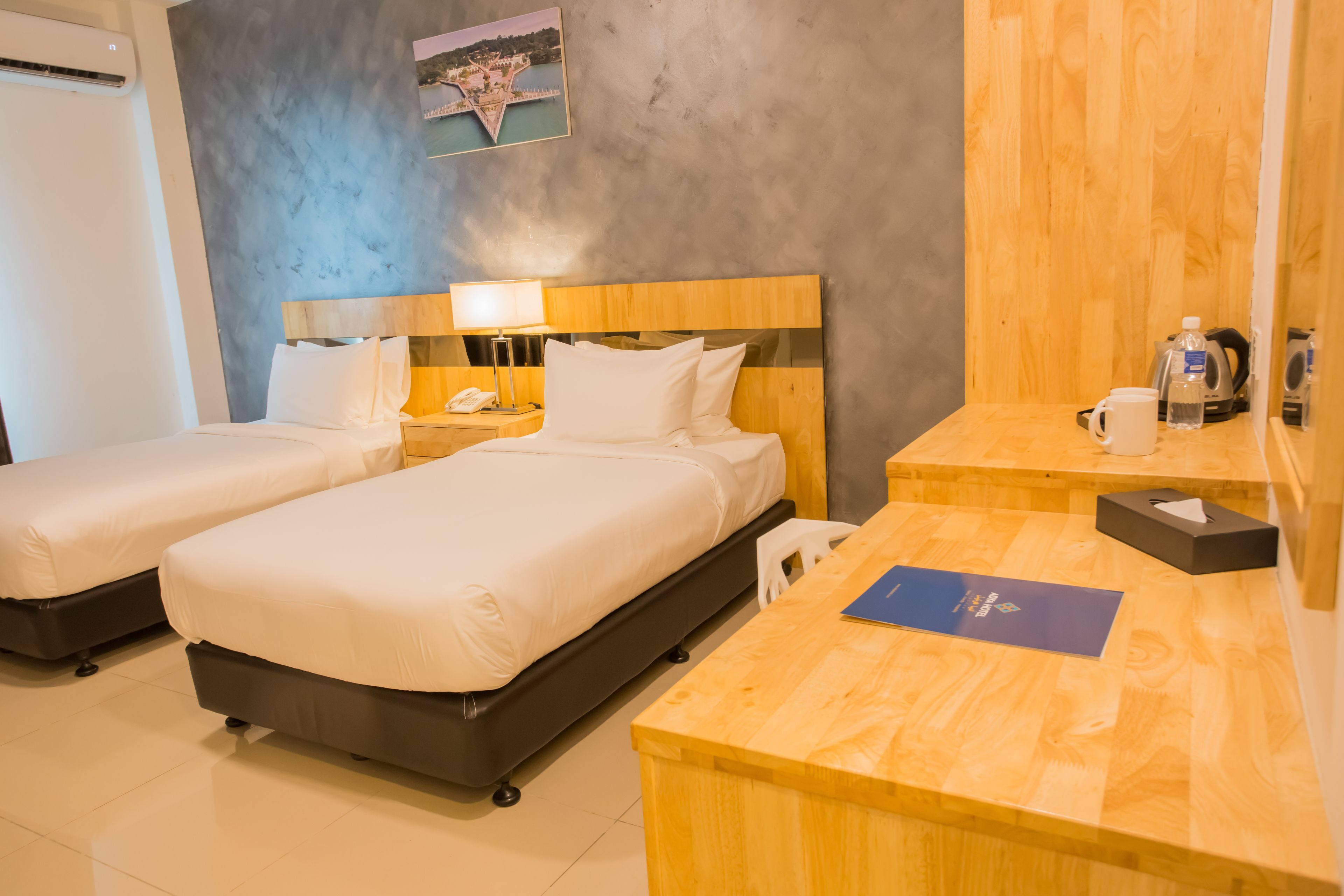 A hotel room with two beds, a wooden desk, a nightstand, a lamp, a phone, a water bottle, a coffee maker, and a framed picture on the wall.