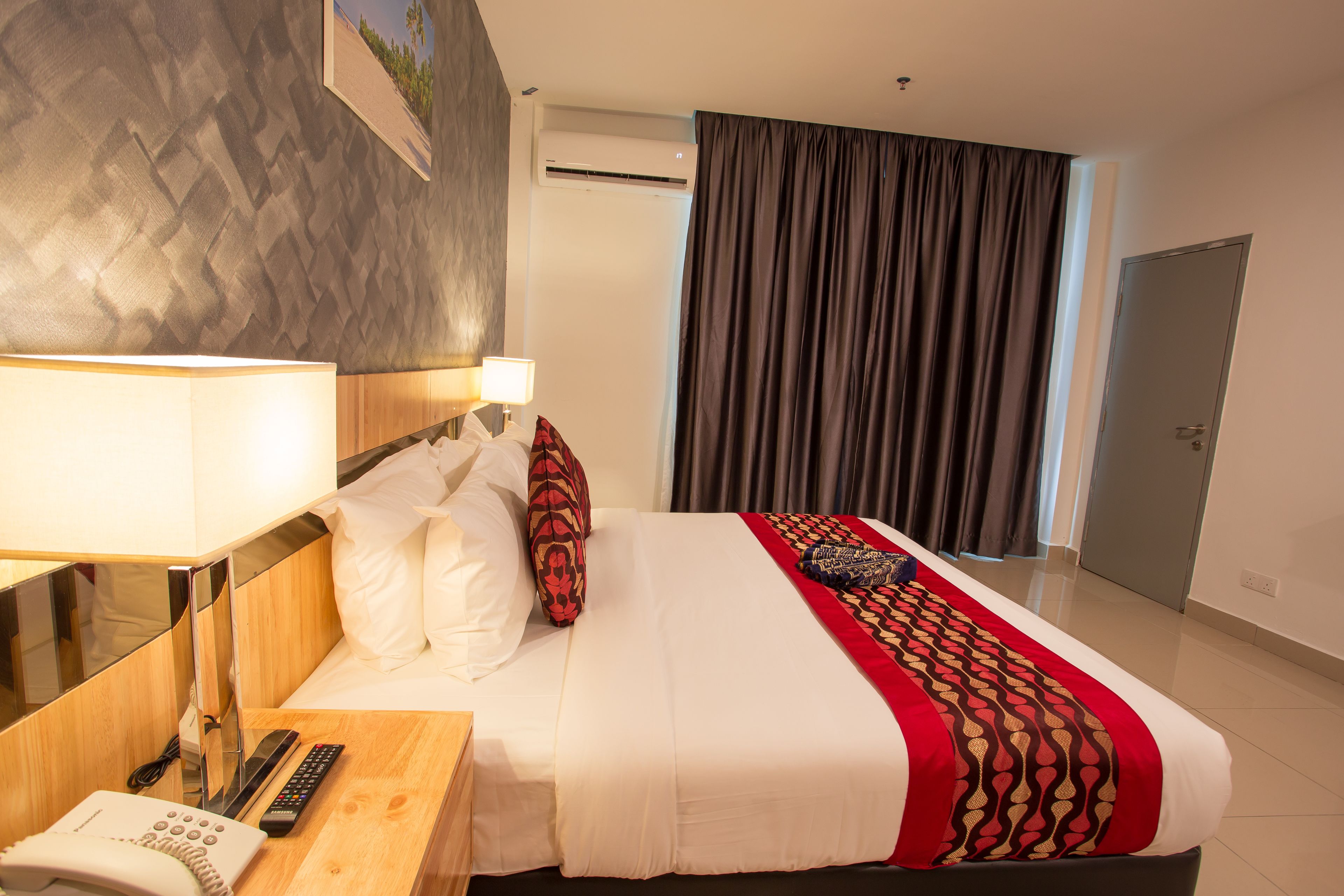A hotel room with a bed, a bedside table with a lamp, a telephone, and a wall-mounted air conditioner.