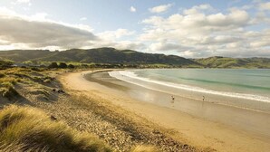 Une plage dont le rivage est incurvé, bordée de dunes herbeuses, avec une colline visible au loin.