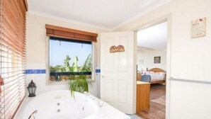 Une salle de bain avec une grande baignoire, une fenêtre avec des stores et une porte donnant sur une autre pièce.