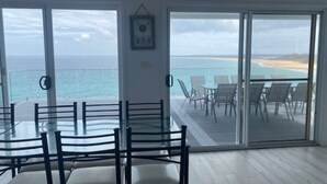 Una habitación con una mesa y sillas de vidrio, una puerta corrediza de vidrio que da a una terraza con vistas a la playa.