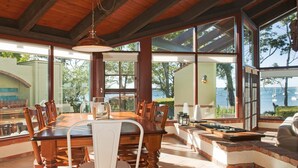 A spacious dining area with a wooden table and chairs, large windows offering a view of the sea, and a cozy seating area with cushions. - Izba - on the foreshore of Callala Bay (Callala Bay)
