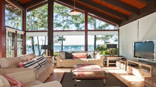 A spacious living room with large windows overlooking a marina, furnished with sofas and a television. - Izba - on the foreshore of Callala Bay (Callala Bay)