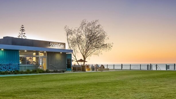 Ett kafé vid stranden med modern design, beläget nära vattnet, med en gräsyta och ett träd.