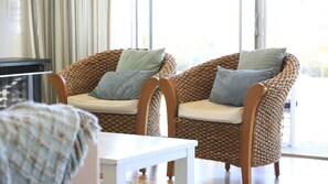 A room with two wicker chairs with cushions, a white table, and a window with curtains.