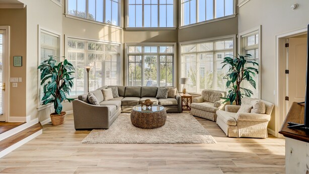 A spacious living room with a large sectional sofa, a round coffee table, and two armchairs. There are large windows providing ample natural light and a view of the outdoors. - 2nd Row luxury home with pool, tiki bar, private beach boardwalk- 30 Knotts Way (Hilton Head Island)