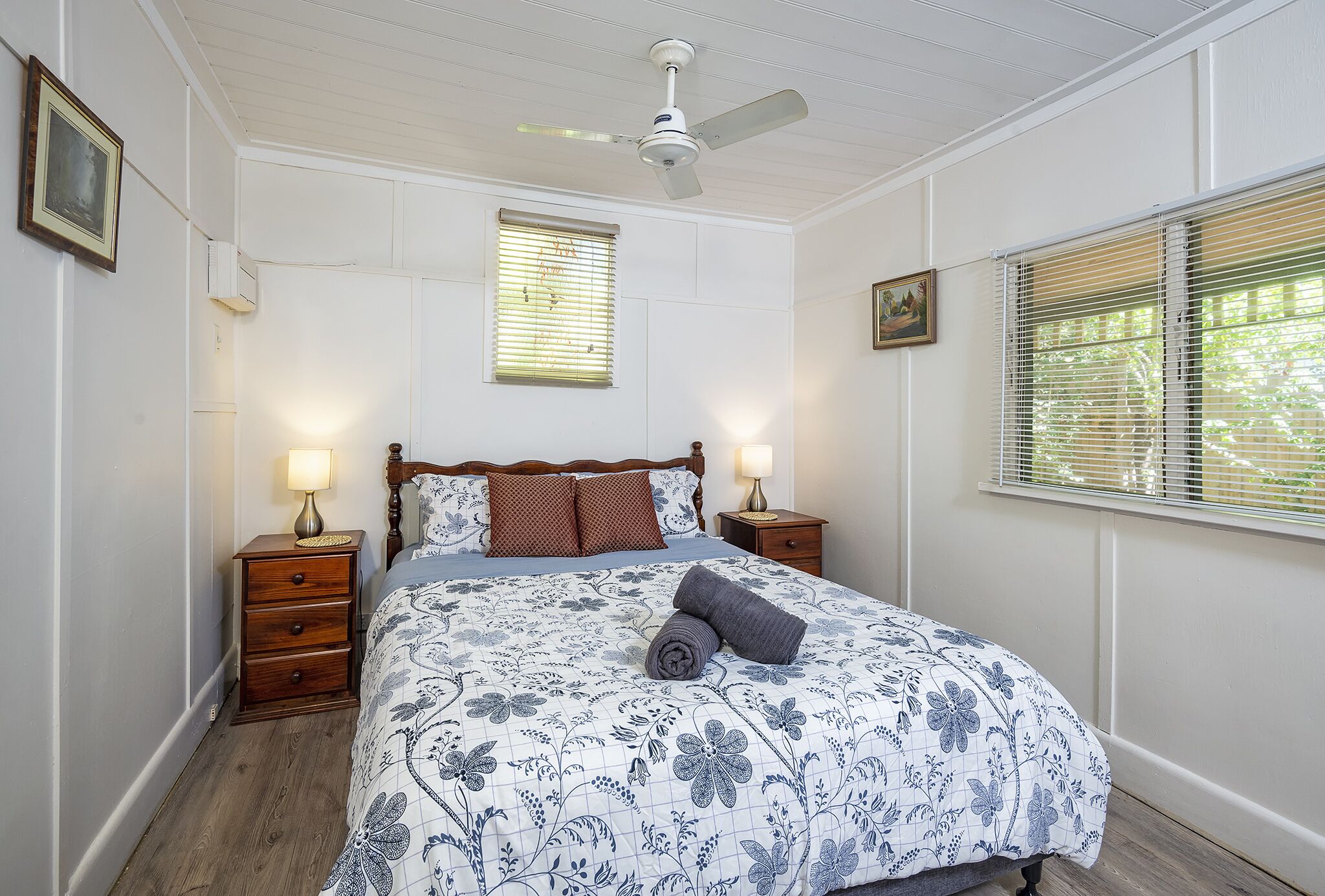 A bedroom with a wooden bed, two bedside tables with lamps, a ceiling fan, and a window with blinds.
