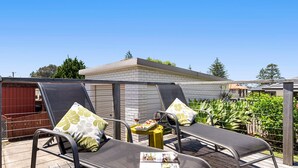 An outdoor patio area with two lounge chairs, a small table with a tray, and a building in the background. - Time To Unwind – cute beach house (Culburra Beach)