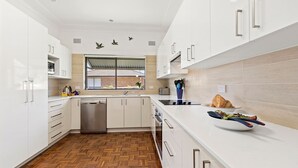 A modern kitchen with white cabinetry, a herringbone wood floor, a built-in dishwasher, and a window with a view of a brick building. - Time To Unwind – cute beach house (Culburra Beach)