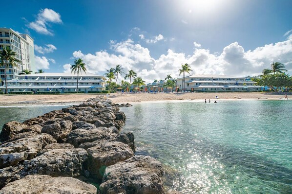 A beachfront hotel with multiple balconies, palm trees, and a rocky shoreline.
