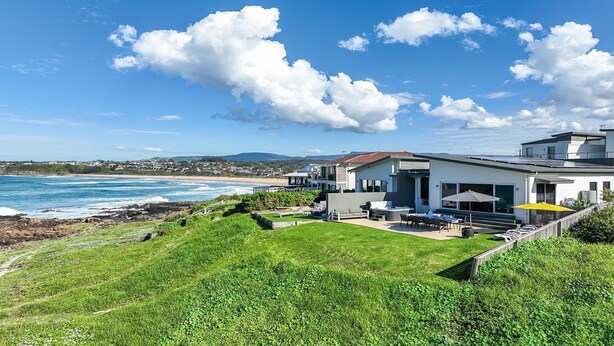 A modern coastal house with a patio overlooking the ocean, a well-maintained lawn, and a clear view of the beach and distant mountains.