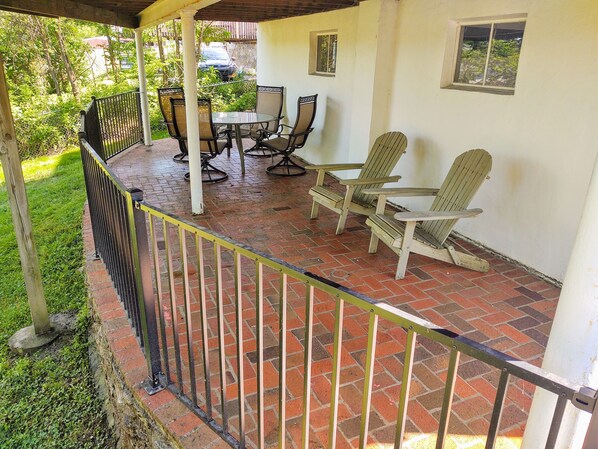 Eine überdachte Terrasse mit Backsteinboden, einem Tisch mit Stühlen und zwei Adirondack-Stühlen.