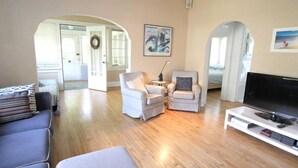 Un salon spacieux avec une grande télévision, deux fauteuils blancs, un canapé et un plancher en bois.
