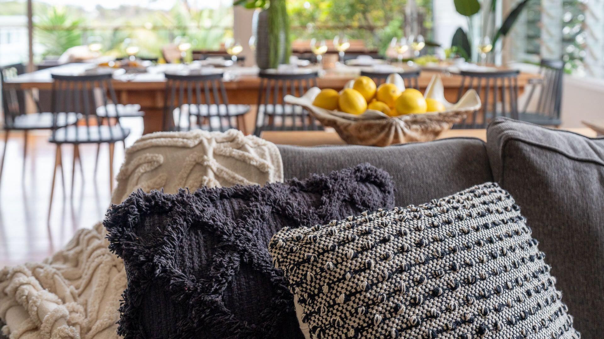 A cozy living room with a grey sofa, a black and white patterned throw pillow, and a beige throw blanket. In the background, there is a dining area with a table set for a meal, a bowl of lemons, and a large potted plant.
