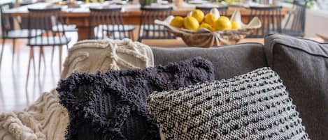 A cozy living room with a grey sofa, a black and white patterned throw pillow, and a beige throw blanket. In the background, there is a dining area with a table set for a meal, a bowl of lemons, and a large potted plant.