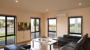Een ruime kamer met grote ramen, een wandgemonteerde flatscreen televisie, een glazen salontafel en zwarte leren banken.