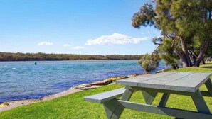 A picnic table by a lake with a grassy area and trees. - Bij's on the Beach – walking distance to beach (Lake Conjola)