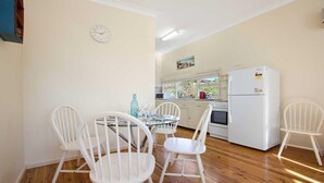 A compact kitchen with white appliances, a dining area with a glass table and white chairs, and a clock on the wall. - Bij's on the Beach – walking distance to beach (Lake Conjola)