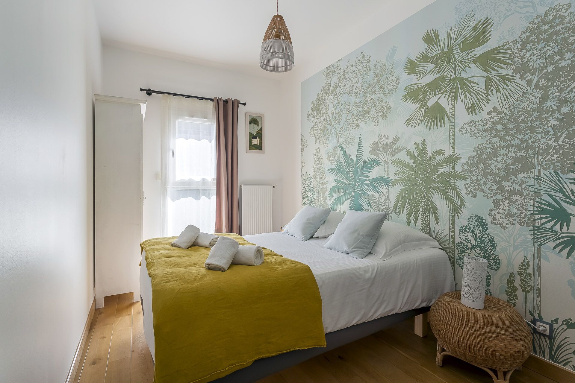 Une chambre avec un lit, une couverture jaune, des oreillers blancs, un luminaire suspendu et un mur orné d’une fresque représentant des plantes tropicales.