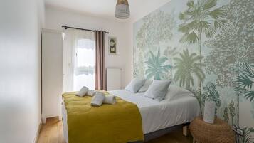 Une chambre avec un lit, une couverture jaune, des oreillers blancs, un luminaire suspendu et un mur orné d’une fresque représentant des plantes tropicales.