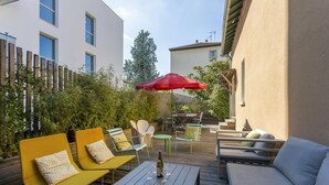 Een terras buiten met houten meubels, waaronder een gele bank en een grijze zithoek, met een rode parasol voor schaduw.
